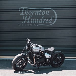 A picture of a Thornton Hundred Motorcycle outside a garage sat in the sun with the Thornton Hundred Motorcycle logo on the wall in the background